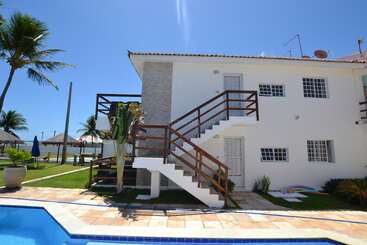A two-story white house with wooden railings, poolside, surrounded by palm trees. Bright sunny day, blue sky, green lawn, patio furniture, and vibrant outdoor atmosphere.