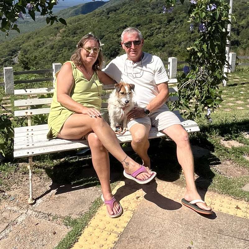 A couple sits closely on a white bench with their small dog, surrounded by lush greenery and hills, enjoying a sunny day outdoors in nature.