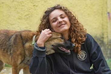 Uma jovem de cabelos cacheados sorri alegremente, com os olhos fechados, enquanto abraça um cachorro marrom todo feliz. O fundo é uma parede amarela, criando uma atmosfera acolhedora e alegre.