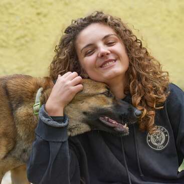 Une jeune femme aux cheveux bouclés sourit joyeusement, les yeux fermés, en serrant dans ses bras un chien brun tout content. En arrière-plan, un mur jaune crée une atmosphère chaleureuse et gaie.