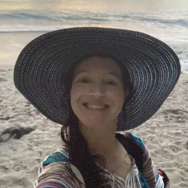 A smiling woman wearing a large, wide-brimmed hat takes a selfie on a sandy beach at sunset, with gentle waves and a colorful sky behind her.
