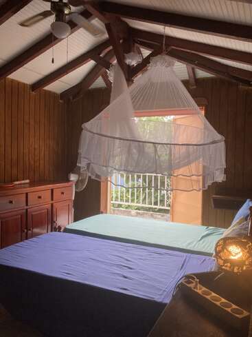 A cozy wooden room with two beds, colorful sheets, a ceiling fan, mosquito net, soft lamp, and an open door letting in sunlight and greenery outside.