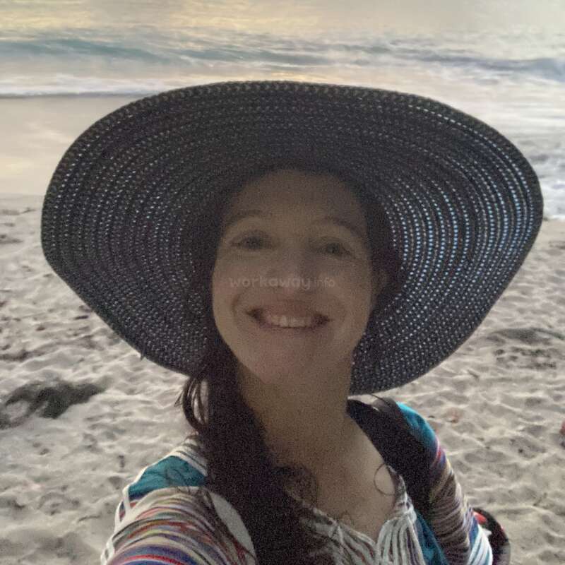 A smiling woman wearing a large, wide-brimmed hat takes a selfie on a sandy beach at sunset, with gentle waves and a colorful sky behind her.