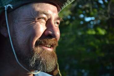 Ein bärtiger Mann, der einen Hut trägt, lächelt strahlend in das warme Sonnenlicht. Um seine Augen herum sind Falten zu sehen. Grünes Laub und verschwommene Bäume bilden den Hintergrund.