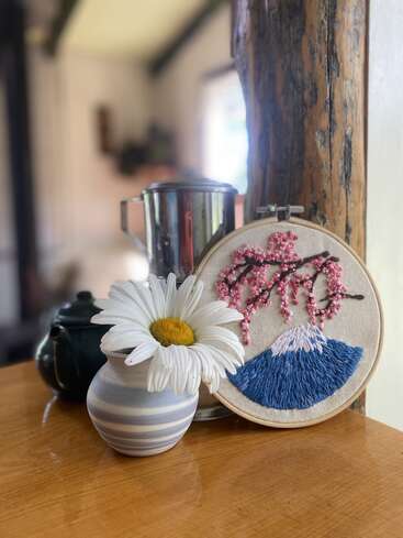 Eine gestreifte Vase mit einem weißen Gänseblümchen, ein bestickter Reifen, der den Berg Fuji und Kirschblüten zeigt, ein Metallkanister und eine Teekanne stehen auf einer Holzfläche.