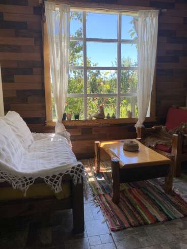 Das Sonnenlicht fällt durch ein großes Fenster mit durchsichtigen Vorhängen und beleuchtet rustikale Holzmöbel, ein gemütliches weißes Sofa, einen bunten Teppich und Topfpflanzen auf der Fensterbank. Friedliches Ambiente.