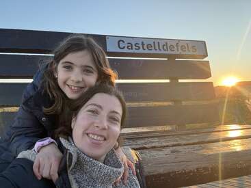 A smiling woman and girl pose for a selfie on a bench at sunset. The sign behind them reads "Castelldefels." Warm, joyful, and peaceful atmosphere.