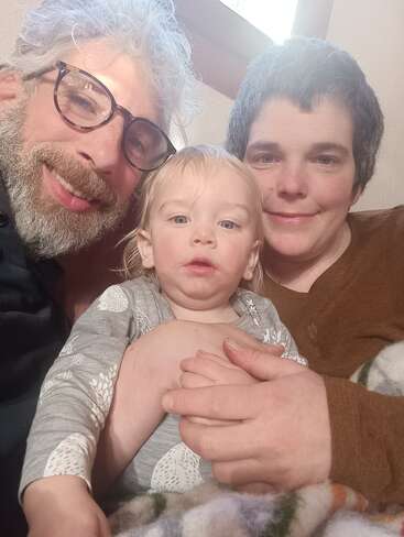 Uma família feliz está sentada bem juntinha, sorrindo para uma selfie. A criança está sentada entre dois adultos, banhada por uma luz natural suave, transmitindo calor humano e união.