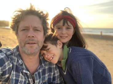The image shows a man and two children posing for a selfie on a beach, with the sun shining brightly behind them.