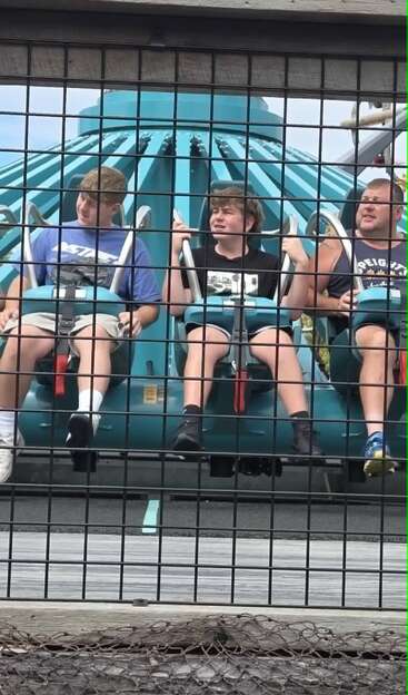 Three people sit on an amusement park ride, secured with harnesses behind a black metal fence. They appear to be waiting, displaying a mix of anticipation and nervousness.