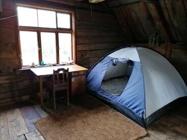 A cozy rustic room with wooden walls and floors contains a tent, a small desk with a chair by a window, and some scattered items on the desk.