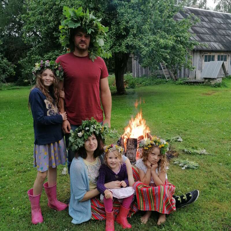 A family, wearing flower and leaf crowns, poses outdoors around a fire. They're dressed in colorful, festive clothing, with green grass, trees, and a rustic house behind.