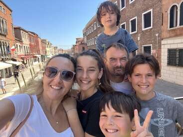 A happy family of six poses for a selfie on a sunny day, surrounded by beautiful old buildings on a charming, colorful European street.