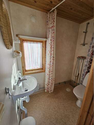 Este acogedor cuarto de baño cuenta con un lavabo pequeño, un inodoro, una ducha a ras de suelo con cortina, luz natural que entra por una ventana y detalles de madera en el techo.