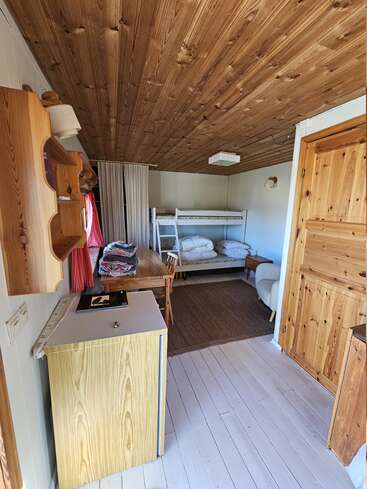 Esta acogedora habitación tipo cabaña de madera cuenta con una litera, una pequeña mesa de comedor con manteles cuidadosamente doblados, una mininevera, estanterías, paneles de madera clara y una decoración sencilla y rústica.