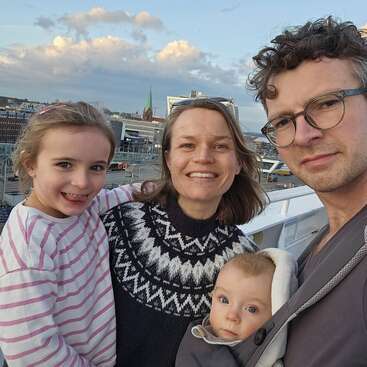 Una alegre familia de cuatro miembros posa al aire libre, sonriendo a la cámara. Al fondo se ve un paisaje urbano con edificios, una iglesia, nubes y un cielo azul.