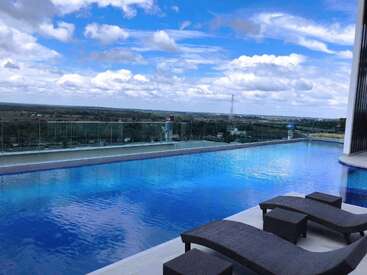 Una serena piscina infinita en la azotea, con lujosas tumbonas, domina un amplio paisaje. El agua refleja el cielo azul brillante lleno de nubes esponjosas y dispersas.