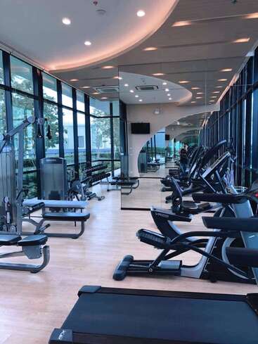 Este moderno gimnasio cuenta con cintas de correr, máquinas elípticas, equipos de fuerza, espejos, grandes ventanales con vistas al exterior, suelo de madera y lámparas de techo, lo que crea un entorno de entrenamiento amplio y bien iluminado.