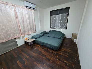Esta habitación cuenta con una cama baja con ropa de cama verde, suelo de madera, muebles mínimos, cortinas rosas, una pequeña mesilla de noche con objetos y aire acondicionado.