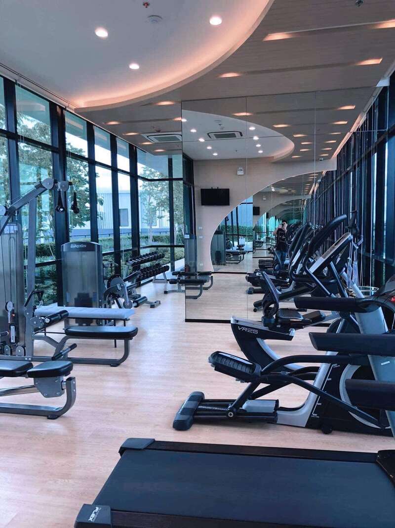 Este moderno gimnasio cuenta con cintas de correr, máquinas elípticas, equipos de fuerza, espejos, grandes ventanales con vistas al exterior, suelo de madera y lámparas de techo, lo que crea un entorno de entrenamiento amplio y bien iluminado.