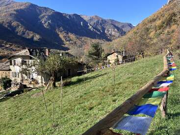 La imagen representa un sereno paisaje montañoso con una casa, árboles y una vibrante valla con una bandera de oración, sobre un cielo azul despejado.