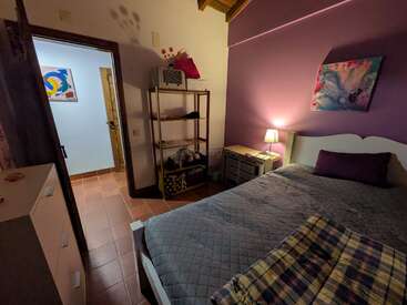 A cozy bedroom with a purple accent wall, bed, bedside table with lamp, open shelf, dresser, and colorful art. Warm lighting creates a relaxing atmosphere.