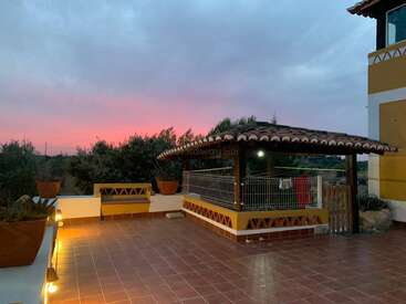 A spacious tiled patio at sunset features a covered pergola, potted plants, benches, and warm lights, creating a cozy outdoor lounge with a beautiful sky.