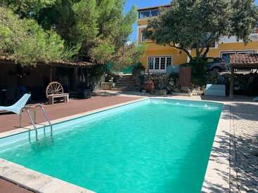 A rectangular swimming pool sits beneath trees, next to a yellow house. There’s a shaded seating area, rustic wagon wheel decor, steps, and a paved courtyard.