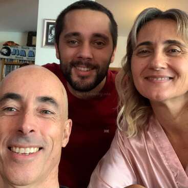 Three adults are smiling for a close-up photo indoors. One man is bald, another has dark hair and beard, and a woman with light hair is beside them.