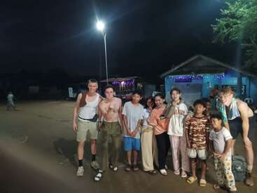 Un grupo de personas, entre ellas adultos y niños, posan juntos de noche bajo una farola, sonrientes y de pie en un camino de tierra de un pueblo.