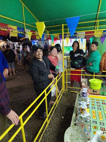 Un grupo de personas se para ante un colorido puesto de feria con barandillas amarillas, tazas y juegos. Sonríen, disfrutando del ambiente festivo bajo un colorido colorín.