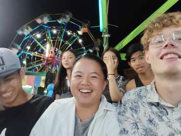 Un grupo de amigos disfruta de una noche en un carnaval, sonriendo y riendo con una noria brillantemente iluminada girando coloridamente en el fondo. Ambiente festivo y alegre.