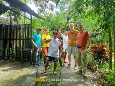 Un grupo de siete hombres posa alegremente al aire libre en un exuberante y verde jardín. Algunos visten ropa informal, otros ropa de trabajo, rodeados de plantas tropicales.