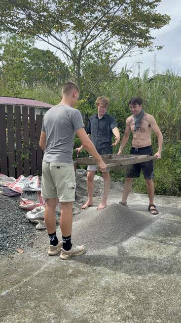Tres jóvenes tamizan arena al aire libre, utilizando un marco de madera. Parecen concentrados, con bolsas de materiales cerca, rodeados de vegetación y árboles bajo un cielo nublado.