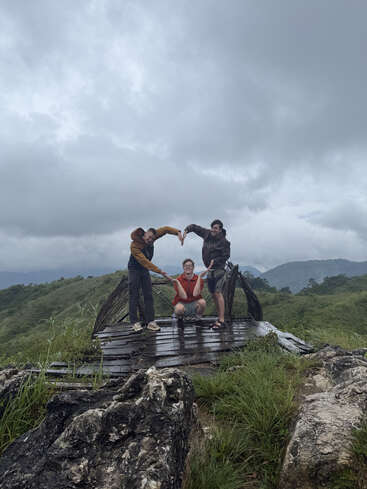 Tres amigos posan creativamente sobre una plataforma de madera en un paisaje verde y exuberante. El cielo nublado se cierne sobre las montañas, capturando juntos un momento de alegría en la naturaleza.