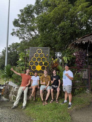 Cinco personas sentadas y de pie frente a un cartel con un diseño de panal, rodeadas de vegetación, sonríen y hacen señas con las manos, con aspecto relajado y feliz.
