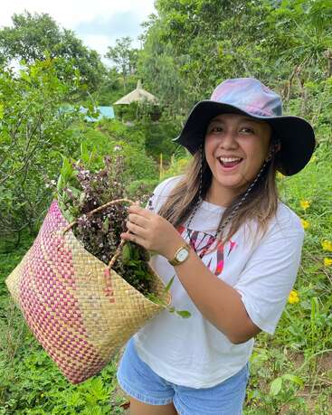 Una mujer sonriente con sombrero de ala ancha y camisa blanca sostiene alegremente una cesta tejida llena de hierbas recién cogidas en un exuberante y verde jardín.