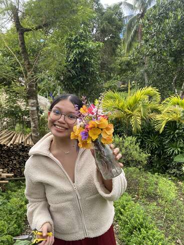 Una chica sonriente con gafas está de pie en un exuberante jardín, sosteniendo un ramo de flores vibrantes en un jarrón de cristal y tijeras de jardinería, mirando alegre.