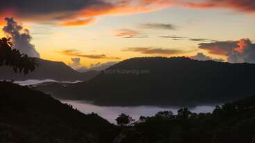 Esta imagen capta un impresionante paisaje de montaña al amanecer o al atardecer, con la niebla llenando los valles, vibrantes colores en el cielo y exuberantes siluetas verdes en primer plano.