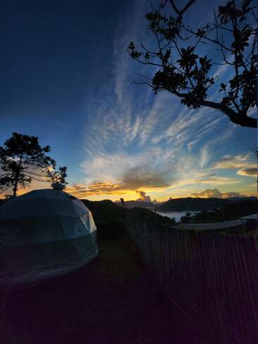 Una impresionante puesta de sol pinta el cielo con vibrantes colores sobre ondulantes colinas. En primer plano, una carpa con forma de cúpula y una valla, mientras los árboles enmarcan el apacible paisaje.