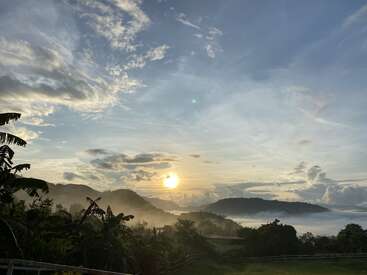 La imagen muestra un hermoso amanecer sobre montañas neblinosas, con exuberante vegetación verde y nubes dispersas en el cielo. Los rayos de sol crean un ambiente sereno y tranquilo.