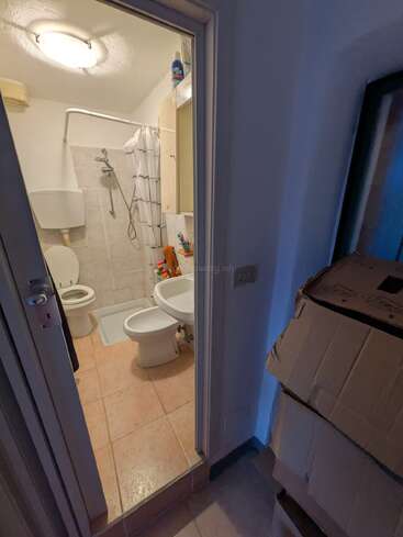 The image shows a small bathroom with a toilet, bidet, sink, and a shower area. Boxes are stacked just outside the bathroom door in a hallway.