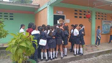 Un groupe d'écoliers en uniforme se rassemble autour d'un adulte à l'extérieur d'une salle de classe et interagit avec enthousiasme. L'arrière-plan montre des murs colorés, des plantes et des affiches éducatives.