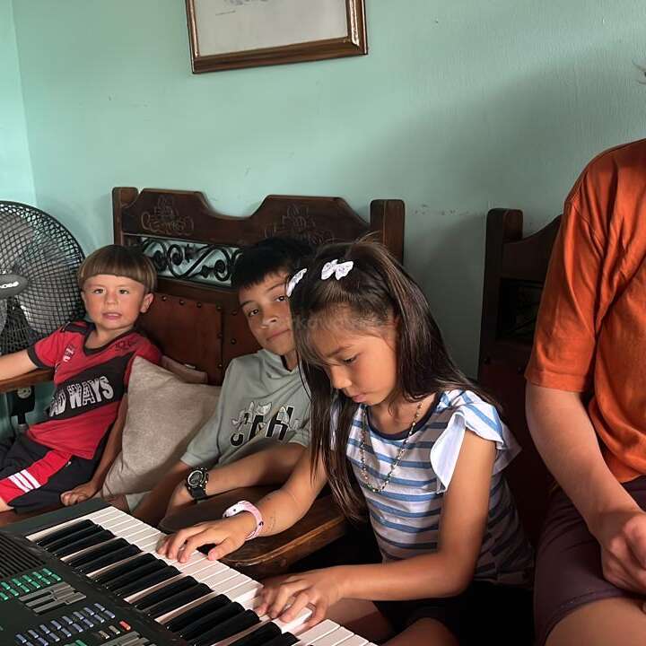 Trois enfants sont assis ensemble à l'intérieur. Une fille joue sur un clavier électrique tandis que deux garçons regardent. Ils ont l'air concentrés et détendus dans un cadre confortable.