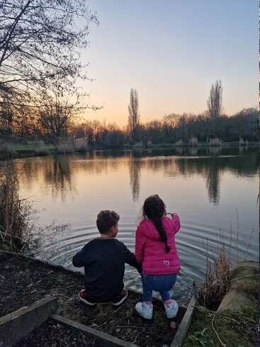 Zwei Kinder sitzen bei Sonnenuntergang an einem stillen See und genießen friedlich die Natur. Das Mädchen zeigt auf das Wasser und verursacht kleine Wellen, umgeben von Bäumen und ruhigen Spiegelungen.