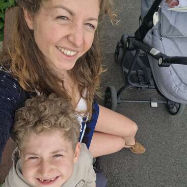 Eine lächelnde Frau und ein kleiner Junge posieren draußen für ein fröhliches Selfie. Neben ihnen steht auf dem Bürgersteig ein Kinderwagen mit einem Baby, was auf einen Familienausflug hindeutet.