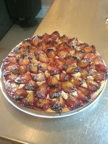 Das Bild zeigt einen runden, goldbraunen Kuchen mit einem Fruchtbelag, der mit Puderzucker bestäubt ist und auf einer weißen Platte auf einem Edelstahltresen steht.