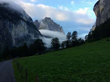 Das Bild zeigt eine ruhige Landschaft mit einer Straße, saftig grünem Gras, Bäumen und majestätischen Bergen, die teilweise von Wolken verdeckt werden.