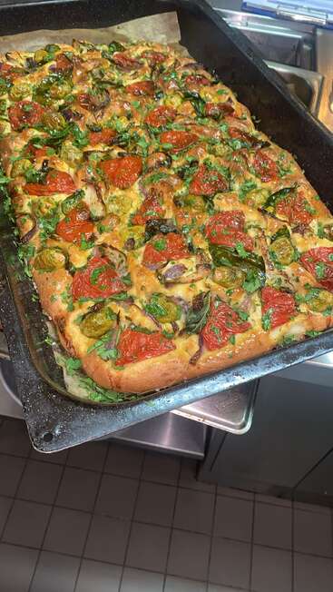 Ein frisch gebackenes Focaccia-Brot liegt auf einem Tablett, belegt mit knackigen Kirschtomaten, grünem Gemüse, Kräutern und Zwiebeln, bereit zum Servieren.