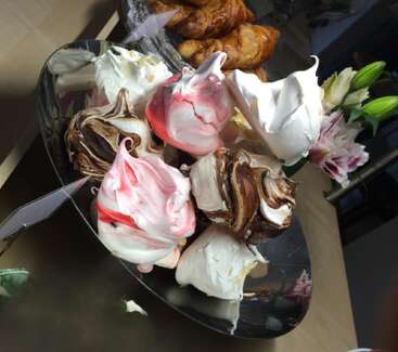 Auf einem schwarzen Teller liegen bunte, luftige Meringues mit rosa, schokoladenbraunen und weißen Strudeln. Im Hintergrund sind Croissants und Blumen zu sehen, die zusammen ein verlockendes Dessertarrangement ergeben.